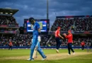 A disappointed Indian batsman walking off the cricket pitch while England players celebrate in the background during a T20 match in a crowded stadium.